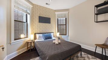 A bedroom with a large bed, two bedside tables, a wall-mounted shelf, and a window with blinds.