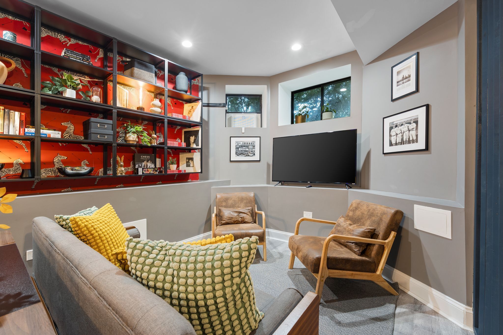 A modern living room with a grey sofa, two brown armchairs, a red bookshelf, a flat-screen TV, and framed pictures on the wall.