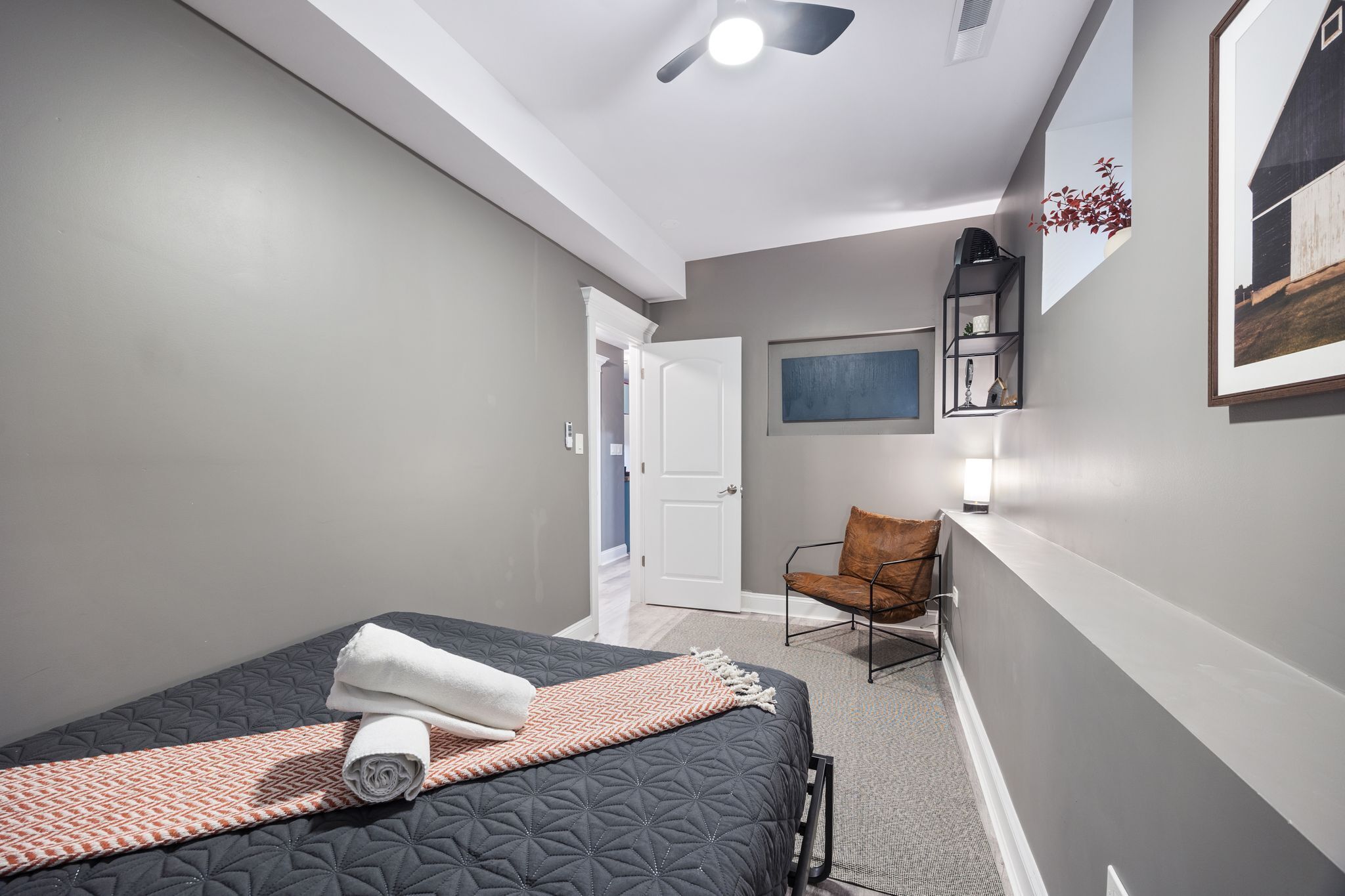 Un dormitorio con cama, una silla, una puerta y una estantería fijada a la pared.