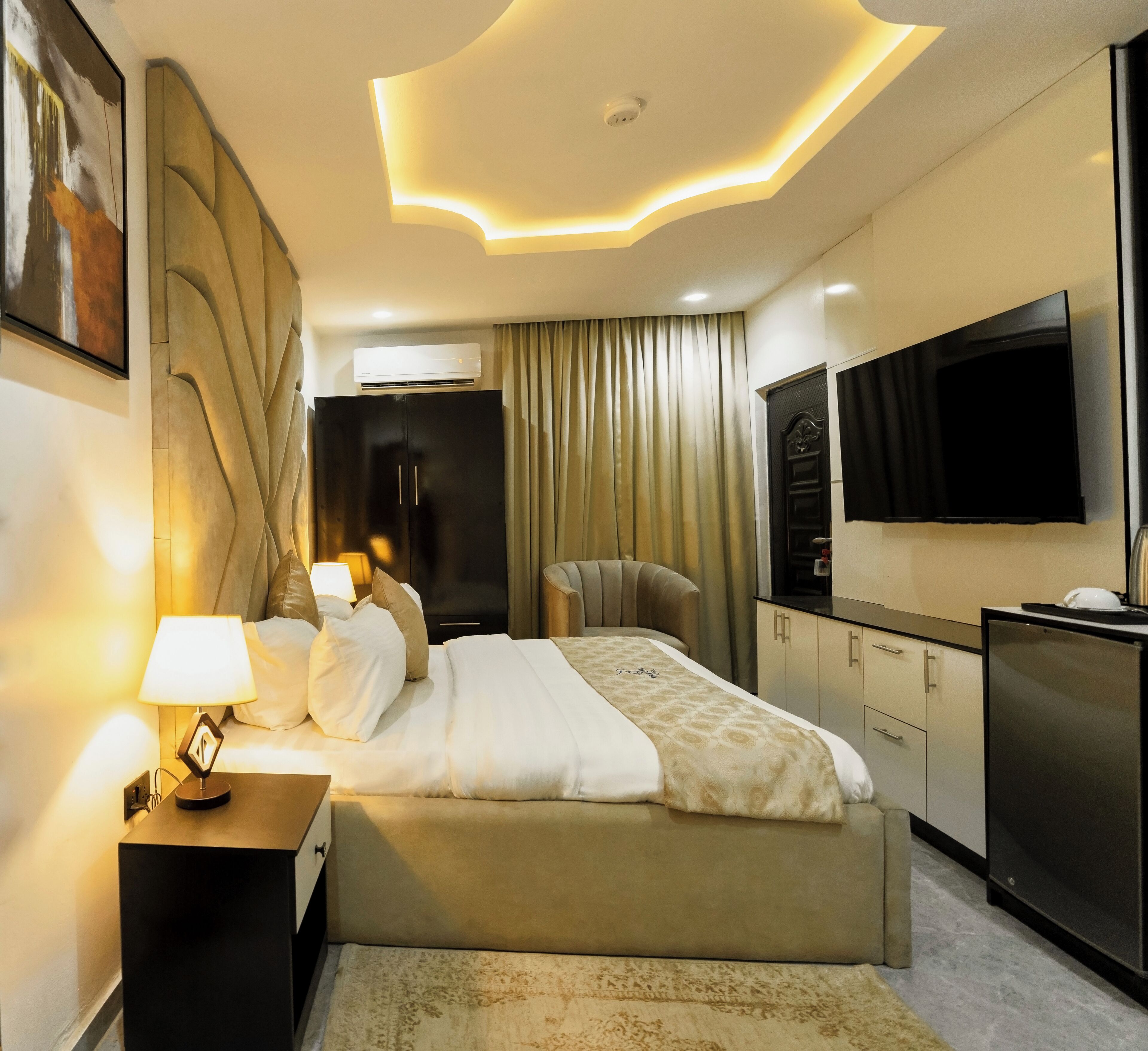 A modern hotel room with a large bed, a flat-screen TV, a minibar, and a ceiling with a unique light design.