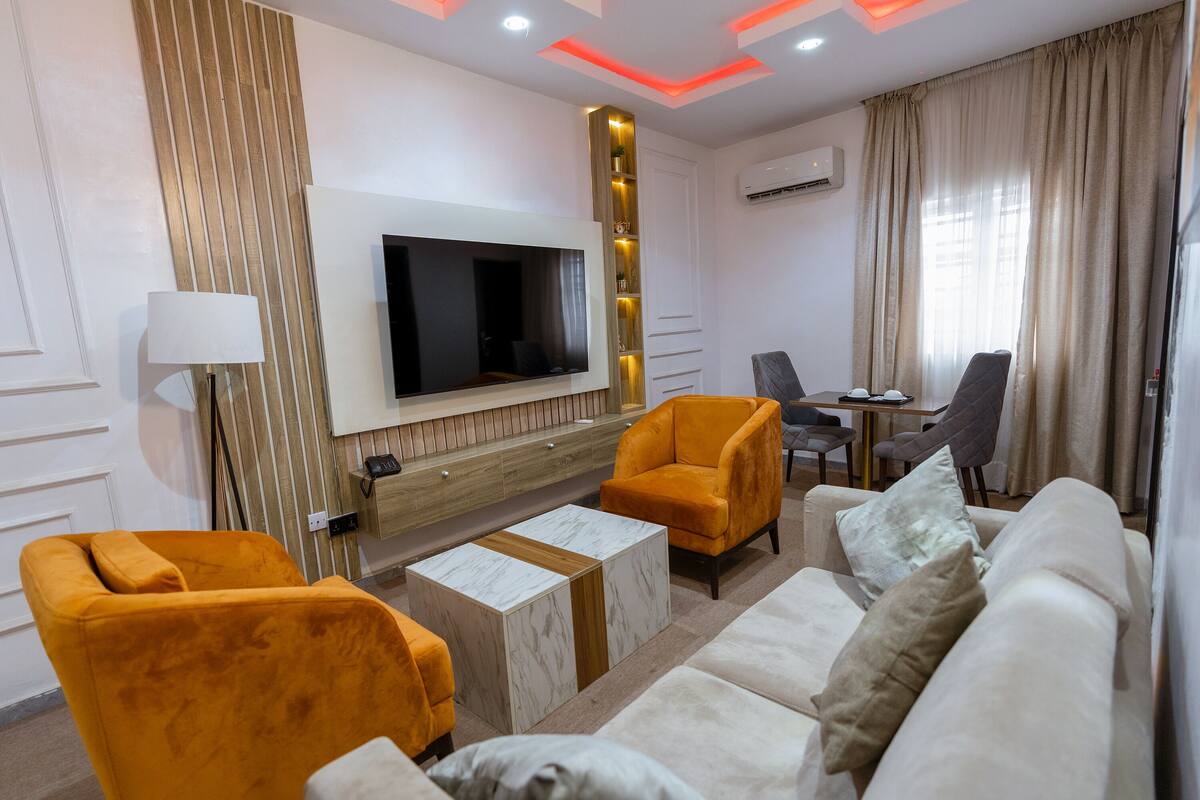 A modern living room with a flat-screen TV, orange armchairs, a beige sofa, and a marble coffee table.