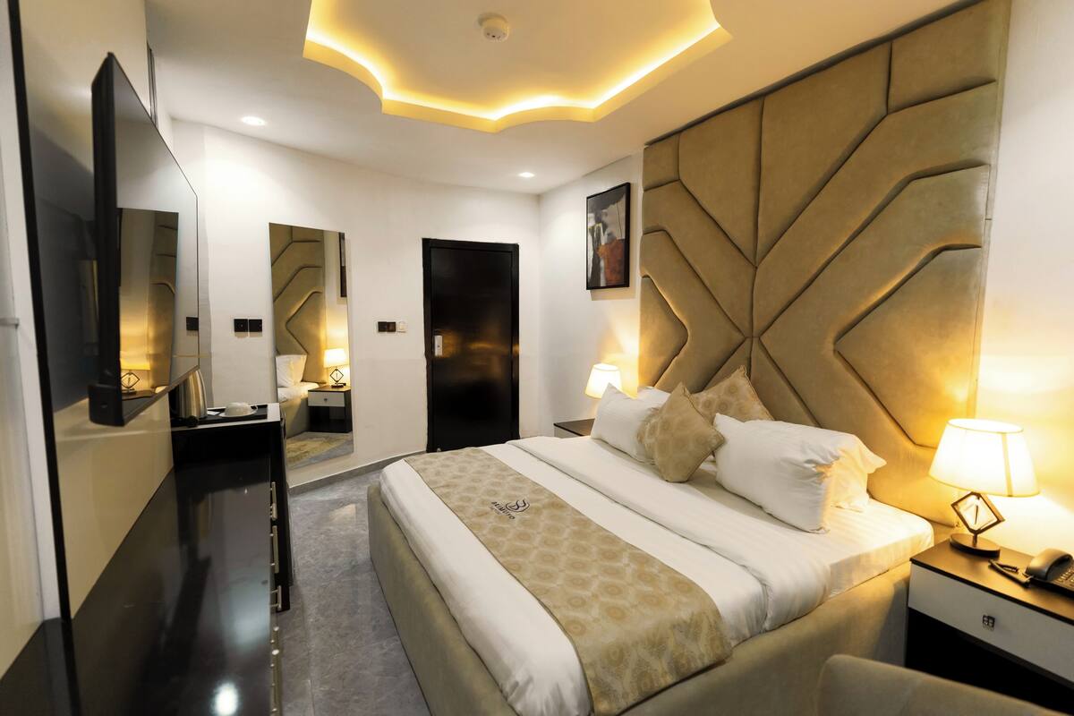 A modern hotel room with a large bed, a headboard with geometric patterns, a bedside table with a lamp, and a mirror on the wall.