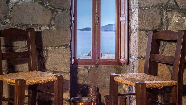 A rustic room with two wooden chairs with woven seats, a stone wall, and a window with a view of the sea and mountains.