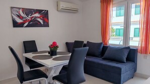 A modern living room with a white dining table, black chairs, a blue sofa with cushions, a painting on the wall, and an air conditioning unit. - Apartments Mihovilovi (Trogir)