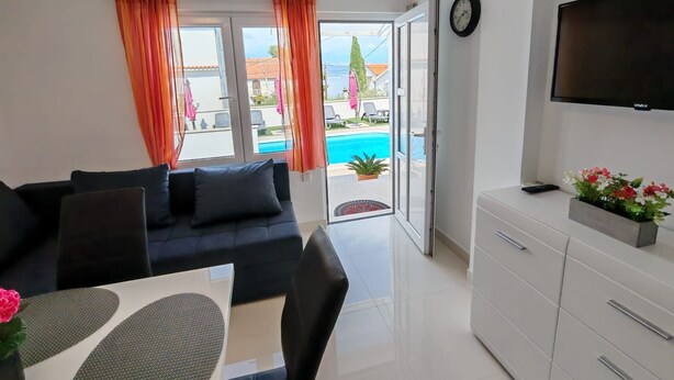 A modern living room with a sofa, a dining table, a TV, and a view of a swimming pool. - Apartments Mihovilovi (Trogir)