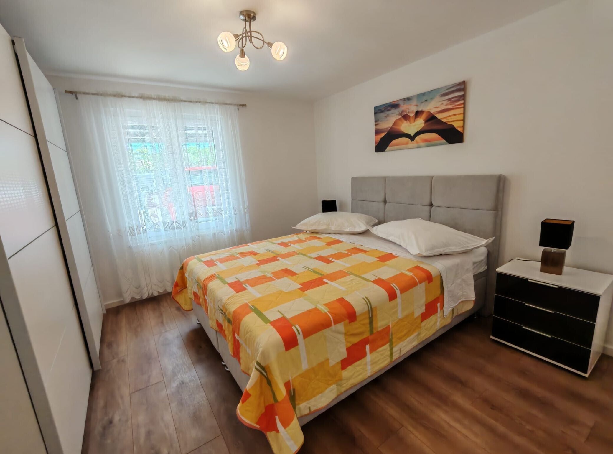 Un dormitorio con cama, mesita de noche, lámpara, un cuadro y una ventana con cortinas.