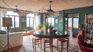 Un comedor con una mesa redonda, sillas y una araña. Se ven ventanas con vistas a zonas verdes y una estantería llena de libros.