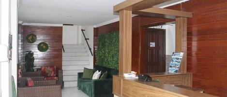 Una zona de recepción con mostrador de madera, un sofá verde, una silla de mimbre y una escalera que conduce a una habitación con una puerta blanca.