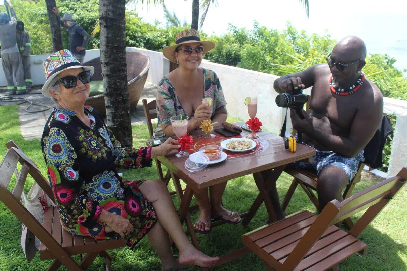 Tres personas sentadas en una mesa de madera al aire libre, con un plato de comida, una bebida y una cámara.