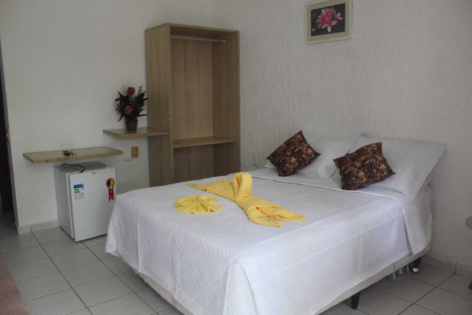 A hotel room with a bed, a small bedside table, a refrigerator, and a wooden wardrobe.