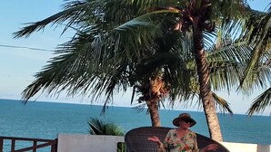 Una mujer sentada en una silla de mimbre junto a una palmera, con vista a una masa de agua.