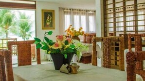 Un comedor con sillas de mimbre, una mesa con un jarrón de flores y vistas a palmeras a través de amplias ventanas.
