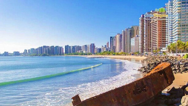 Un paisaje urbano costero con un destacado pecio oxidado en la playa, edificios altos y aguas azul turquesa.