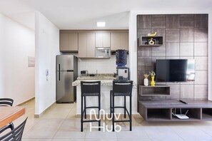 A modern kitchen with a built-in microwave, a refrigerator, and a water dispenser. There is a dining area with two chairs and a table. A flat-screen TV is mounted on a dark wall, and a shelf with decorative items is visible. - Residencial Saint Barth (Ubatuba)
