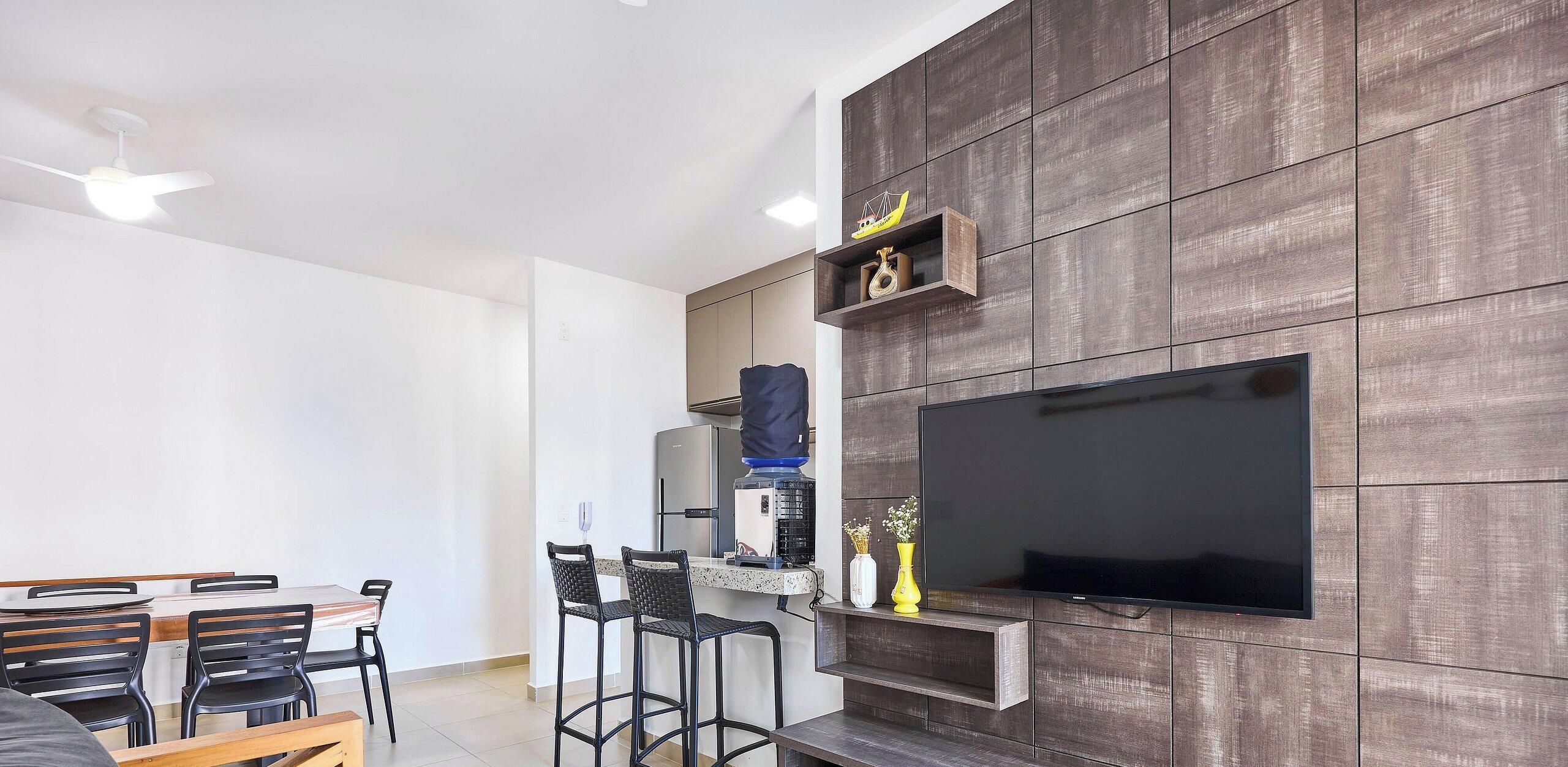 Una sala de estar moderna con un televisor de pantalla plana montado en una pared de madera, un comedor con mesa y sillas, y una cocina con un refrigerador y dispensador de agua.
