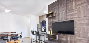 Una sala de estar moderna con un televisor de pantalla plana montado en una pared de madera, un comedor con mesa y sillas, y una cocina con un refrigerador y dispensador de agua.