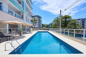 A rectangular swimming pool with a tiled edge, surrounded by a white railing. There are lounge chairs and umbrellas on the poolside. - Residencial Saint Barth (Ubatuba)