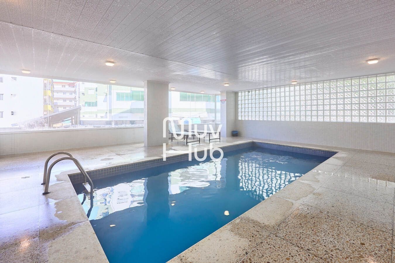 A swimming pool with a tiled floor and a clear blue pool, surrounded by a white tiled wall and a window with a view of buildings outside.