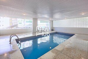 A swimming pool with a tiled floor and a clear blue pool, surrounded by a white tiled wall and a window with a view of buildings outside.