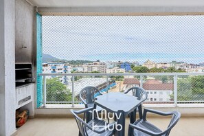 Un balcón con una mesa y sillas, una parrilla, y vistas a una ciudad y montañas.