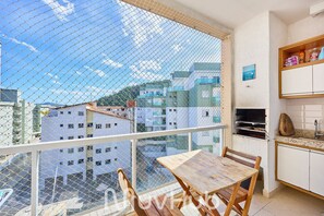 A balcony with a view of buildings, a wooden table, and a kitchen area. - Residencial Saint Barth (Ubatuba)