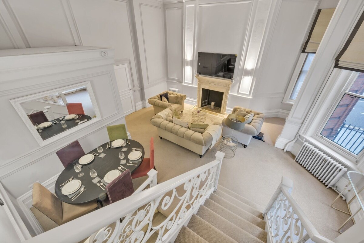 A spacious living area with a fireplace, a dining table set for four, and a staircase leading to an upper floor.