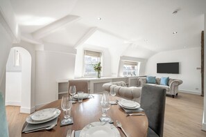 A dining area with a wooden table set for six, a sofa in the background, and a TV mounted on the wall. - The Knightsbridge Residences (London)
