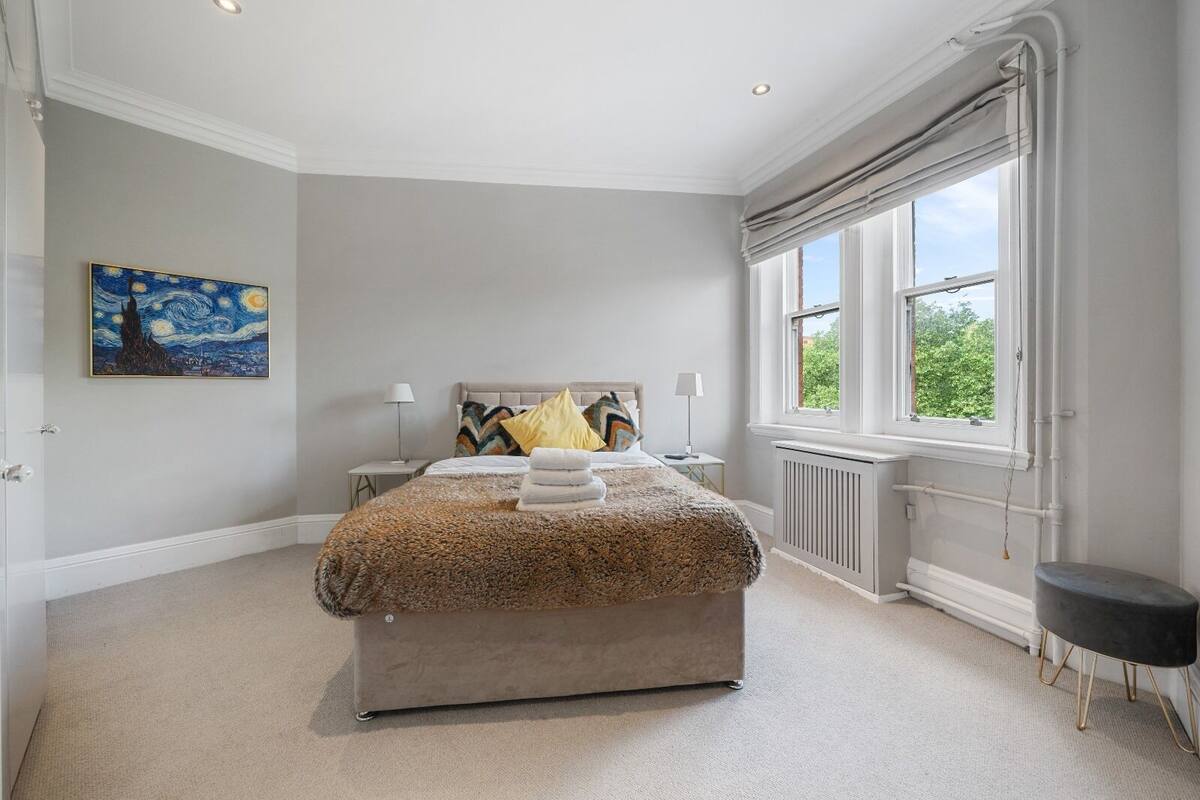 A bedroom with a large bed, a radiator, a window with curtains, a nightstand, a lamp, and a painting on the wall.