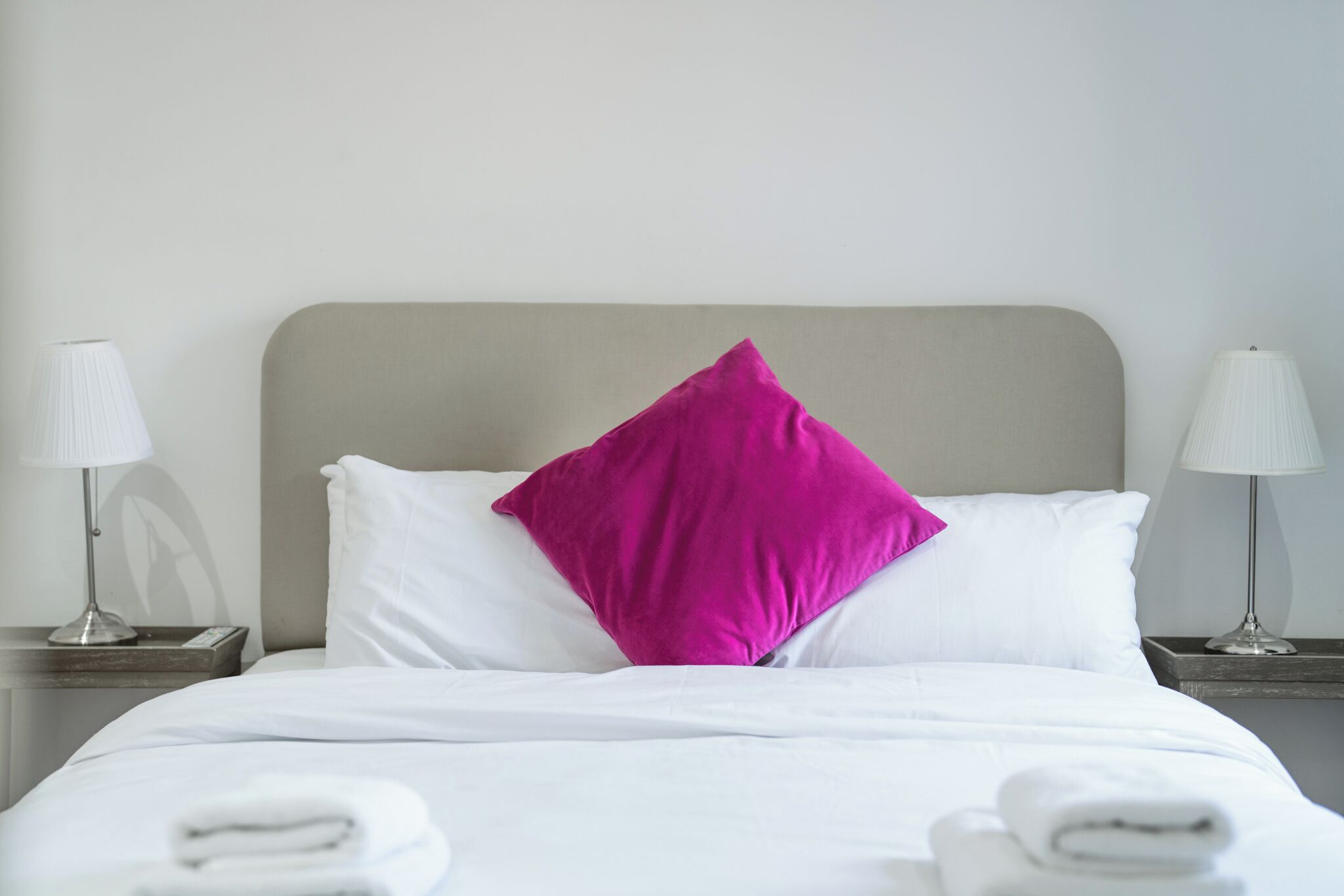 A neatly made bed with white linens and a single bright pink pillow, flanked by two bedside lamps on matching nightstands.
