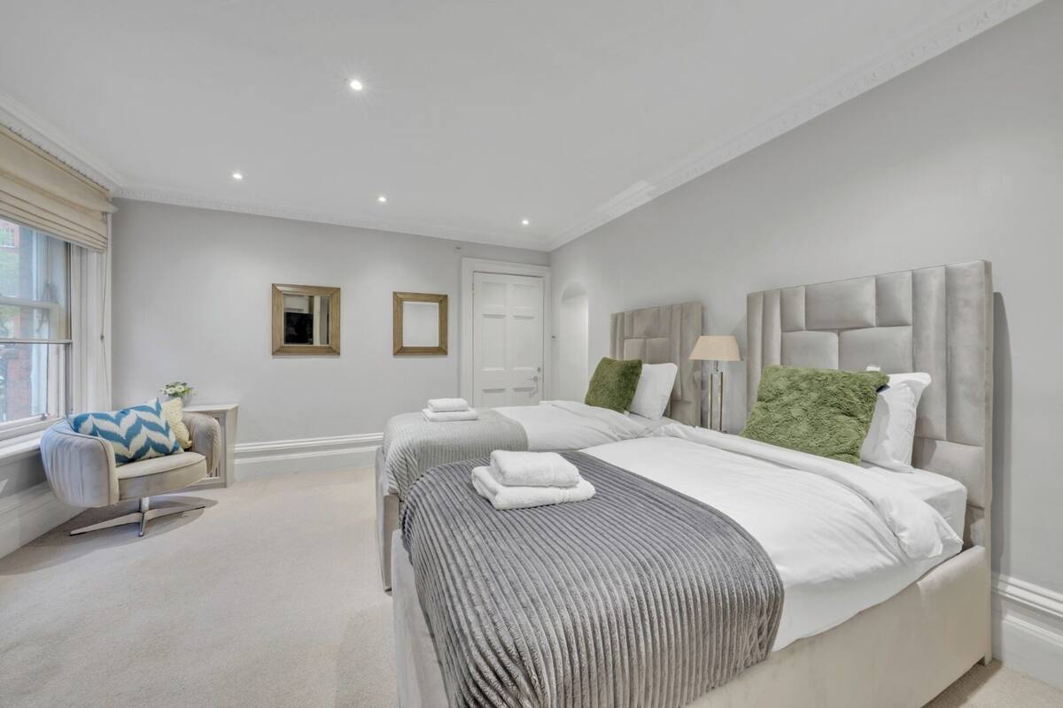 A modern bedroom with two beds, a sitting area with a chair, and a large window.