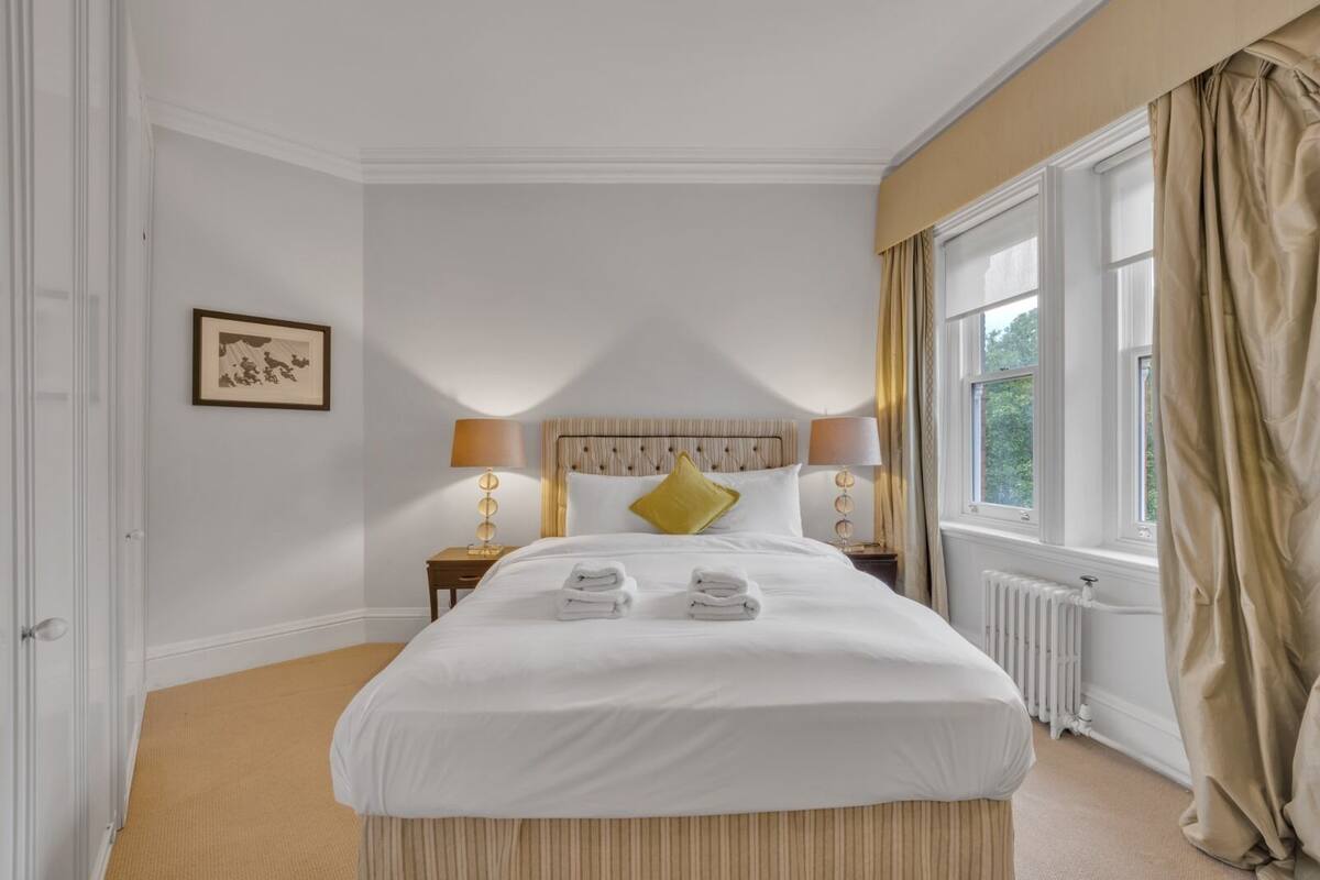 A neatly made bed with folded towels, two bedside lamps, a framed picture on the wall, and a window with curtains.