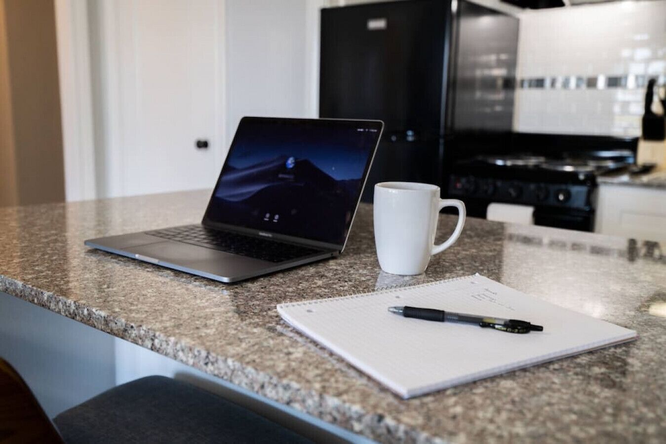 Een keukentafel met een laptop, een witte mok en een notitieboekje met een pen.