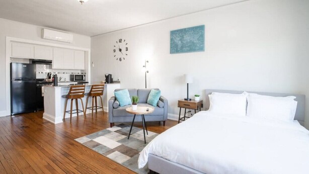 Ein moderner, gut beleuchteter Raum mit einem großen Bett, einem Schlafsofa, einem kleinen Tisch und einer Küchenzeile im Hintergrund.