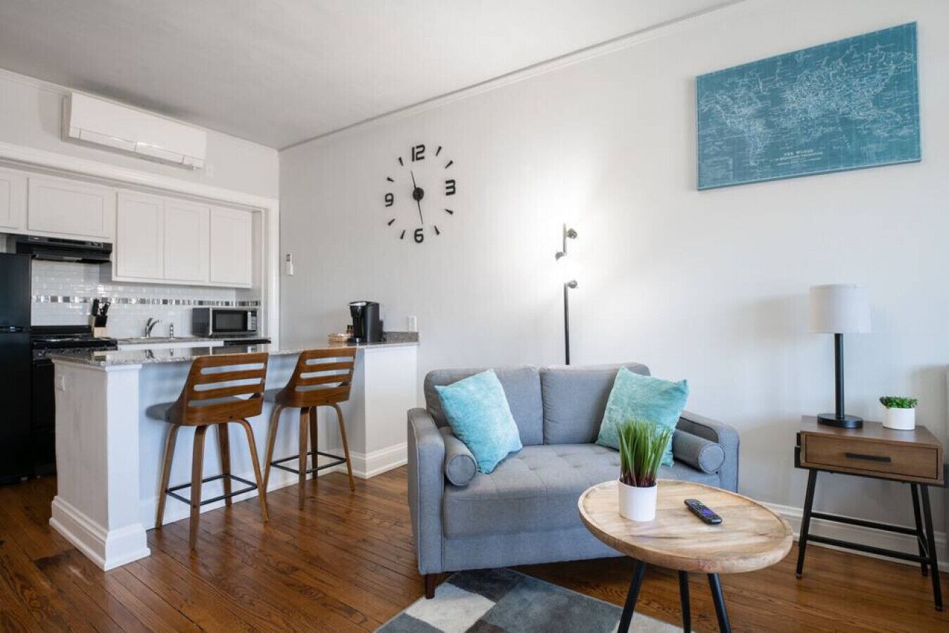 A modern living room with a grey sofa, two wooden bar stools, a round wooden coffee table with a remote control, a small potted plant, a lamp, and a world map on the wall.