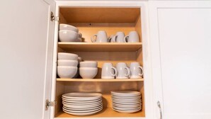 A white cabinet with shelves containing white bowls, mugs, and stacked plates. - WOOD`INN - Cozy 2BD over the bridge (East Peoria)