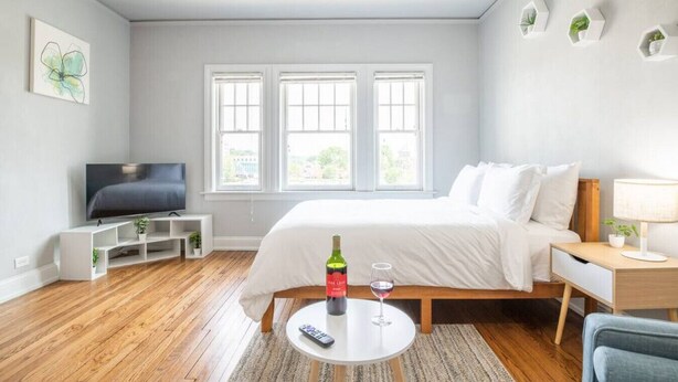 Una habitación de hotel moderna con cama, televisión, mesita de noche con lámpara, una pequeña mesa con botella y vaso, y ventana con vistas.