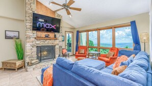A living room with a stone fireplace, a large blue sofa, orange armchairs, and a flat-screen TV mounted on a stone wall.
