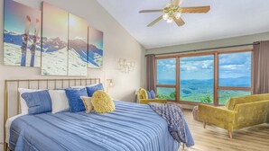 A bedroom with a large bed, a ceiling fan, a view of the mountains through the window, and a yellow armchair.