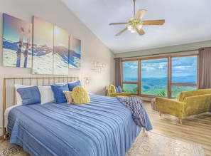 Quarto com uma cama grande, um ventilador de teto, vista para as montanhas pela janela e uma poltrona amarela.