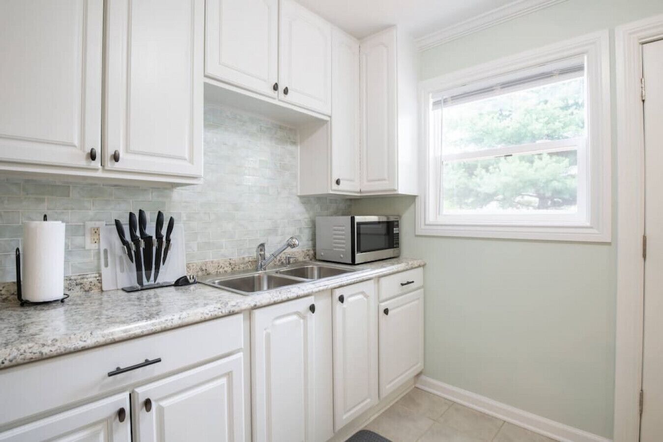 A kitchen with white cabinets, a granite countertop, a sink, a microwave, and a window with a view of greenery.