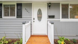 Une maison de plain-pied avec une porte d’entrée blanche, un petit porche doté d’une terrasse en bois et une boîte aux lettres située sur le côté droit.