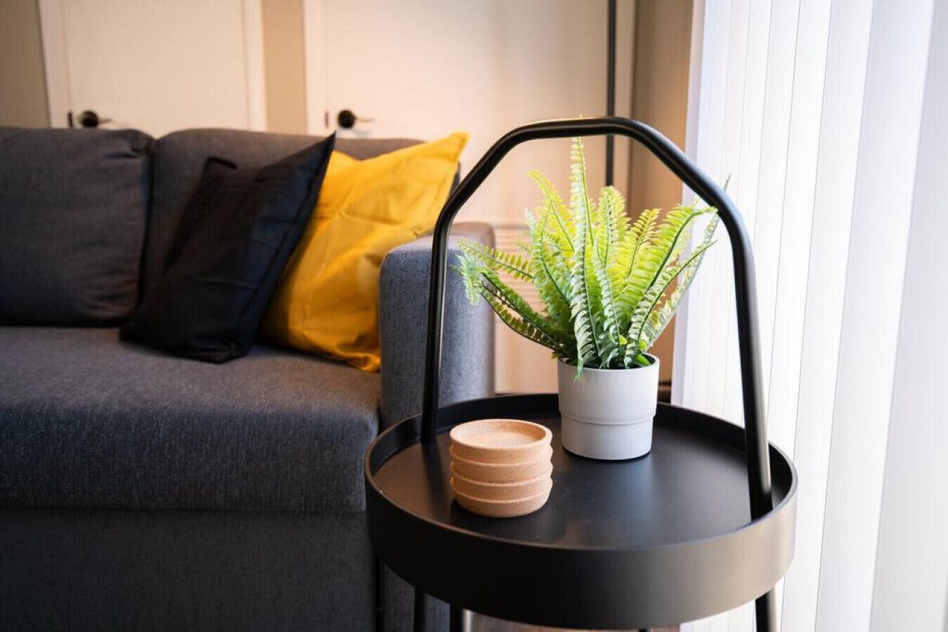 A modern living room with a grey sofa, a yellow pillow, a black side table with a potted fern, and a stack of wooden coasters.