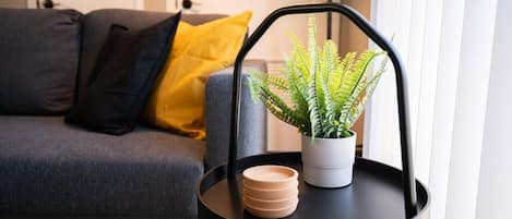A modern living room with a grey sofa, a yellow pillow, a black side table with a potted fern, and a stack of wooden coasters.