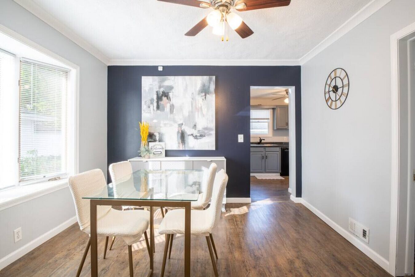 A dining area with a glass table and four chairs, a ceiling fan, and a kitchen visible through an open doorway.