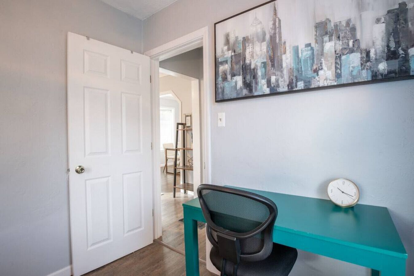 Een kamer met een blauwgroen bureau, een zwarte bureaustoel, een witte deur en een stadsgezicht schilderij aan de muur.
