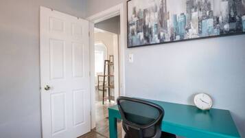 Een kamer met een blauwgroen bureau, een zwarte bureaustoel, een witte deur en een stadsgezicht schilderij aan de muur.