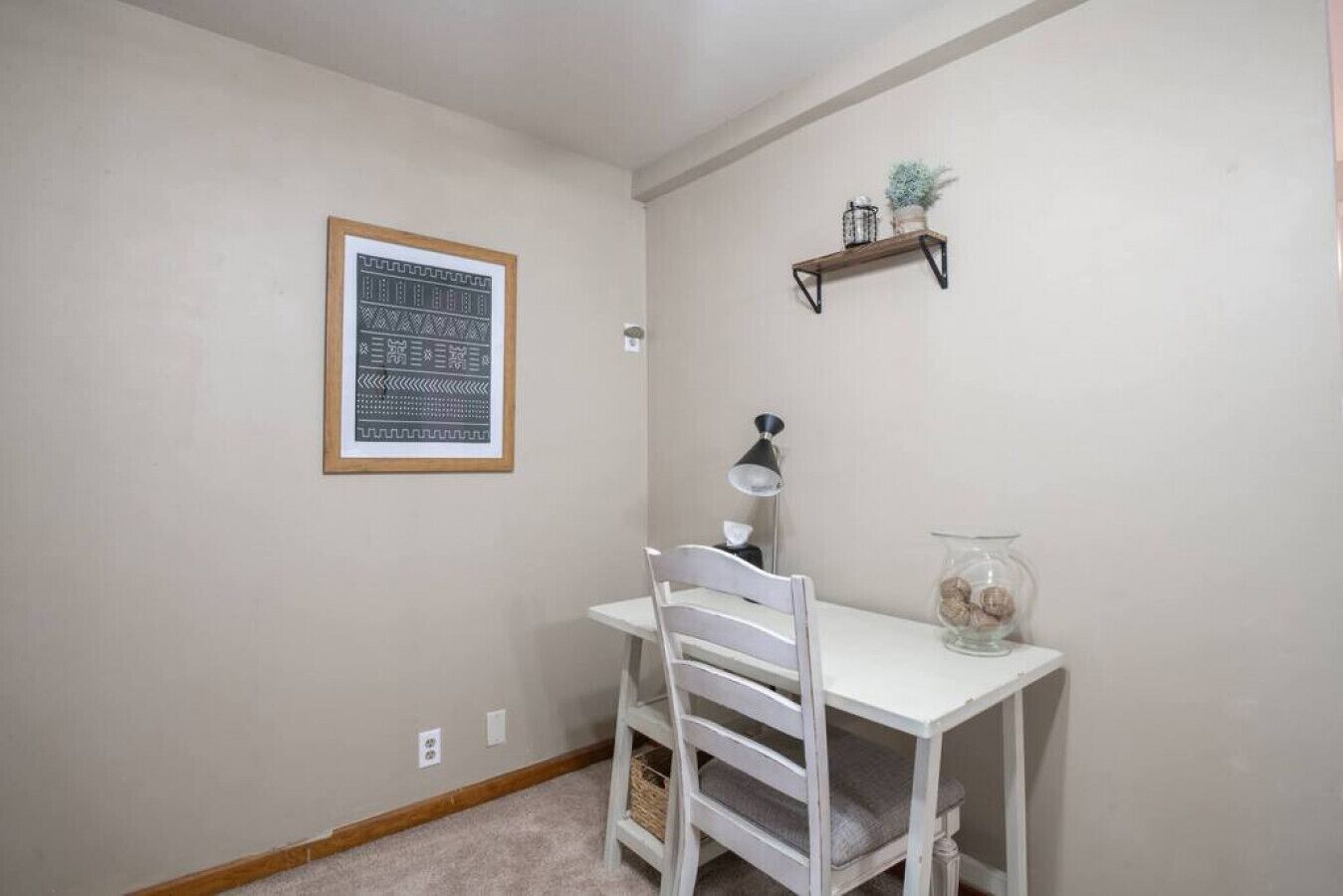 A small, neatly organized study area with a white desk, a chair, a lamp, a shelf with decorative items, and a framed picture on the wall.