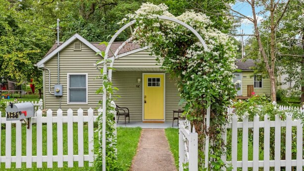 Ein Haus mit gelber Tür, weißem Zäunchen und einem Garten mit weißen blühenden Pflanzen.