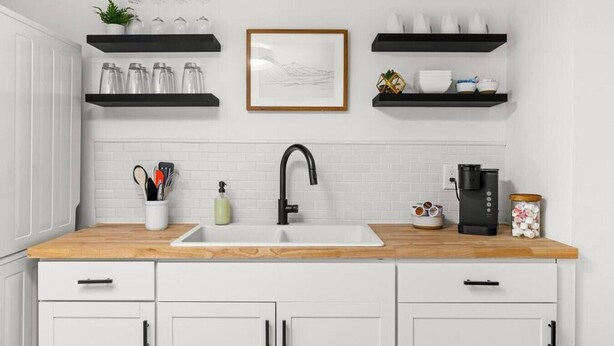A modern kitchen with white cabinets, a wooden countertop, a black faucet, and various kitchen items on shelves. - Halcyon Haven - A Heavenly Homestead for 4 (Morton)