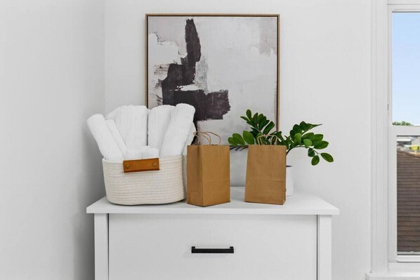 A white cabinet with a framed abstract painting, a woven basket with white towels, and two brown paper bags.